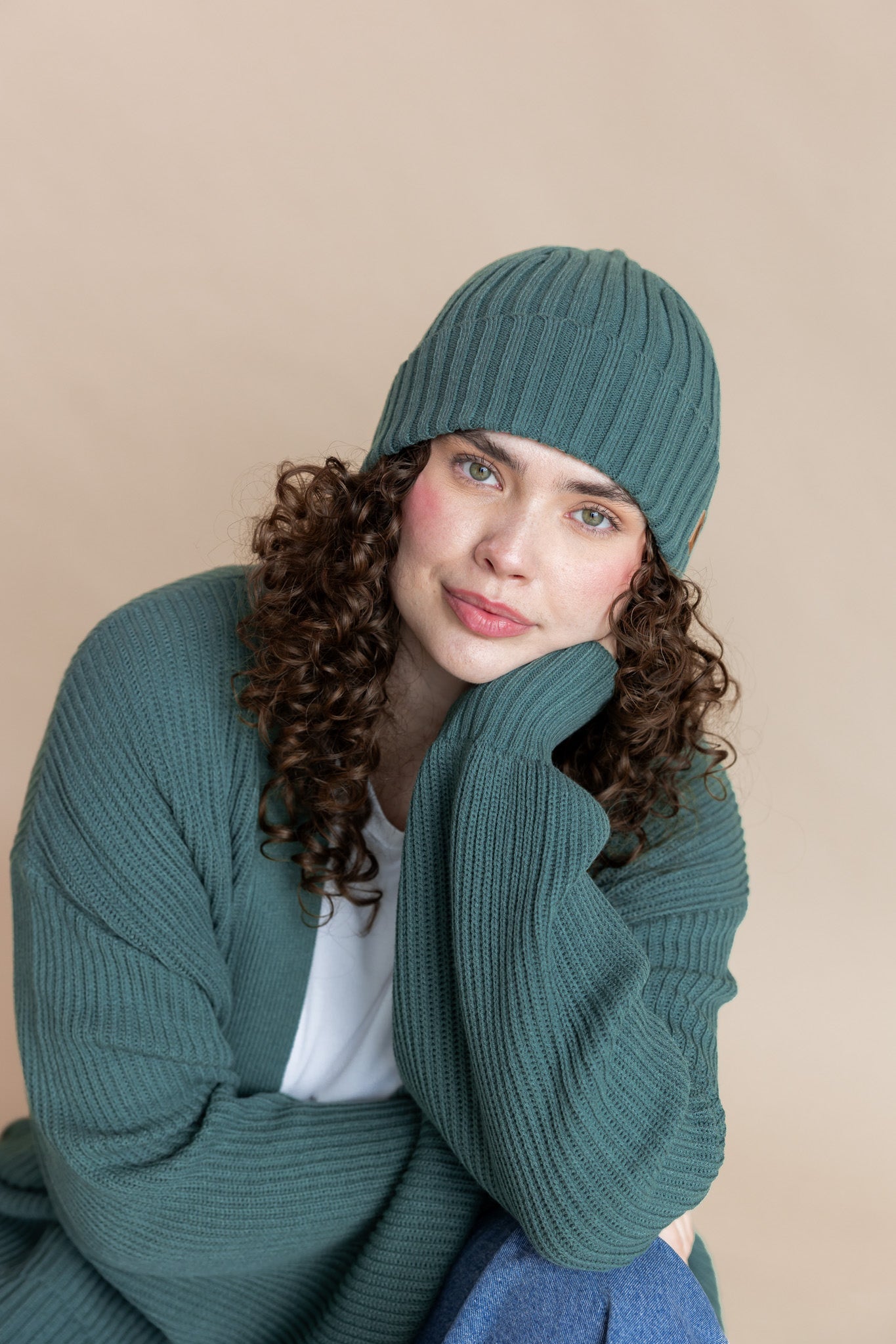 Modal wearing Chunky Knit Adult Ribbed Beanie in pine over white t-shirt and chunky knit women's cardigan in pine and blue jeans on a beige background