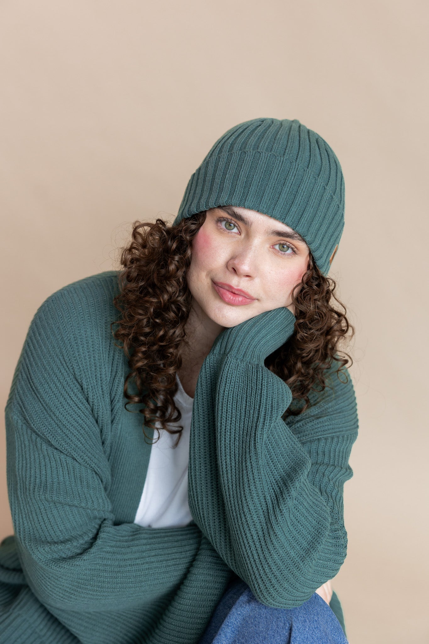 Modal wearing Chunky Knit Adult Ribbed Beanie in pine over white t-shirt and chunky knit women's cardigan in pine and blue jeans on a beige background