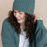 Modal wearing Chunky Knit Adult Ribbed Beanie in pine over white t-shirt and chunky knit women's cardigan in pine on a beige background