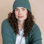 Modal wearing Chunky Knit Adult Ribbed Beanie in pine over white t-shirt and chunky knit women's cardigan in pine on a beige background