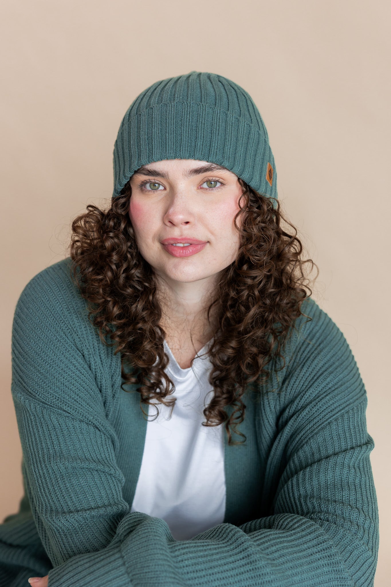 Modal wearing Chunky Knit Adult Ribbed Beanie in pine over white t-shirt and chunky knit women's cardigan in pine on a beige background
