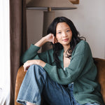 Modal sitting on brown sofa wearing Chunky Knit Women's Waffle Cardigan in Pine over white camisole and blue jeans