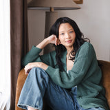 Modal sitting on brown sofa wearing Chunky Knit Women's Waffle Cardigan in Pine over white camisole and blue jeans