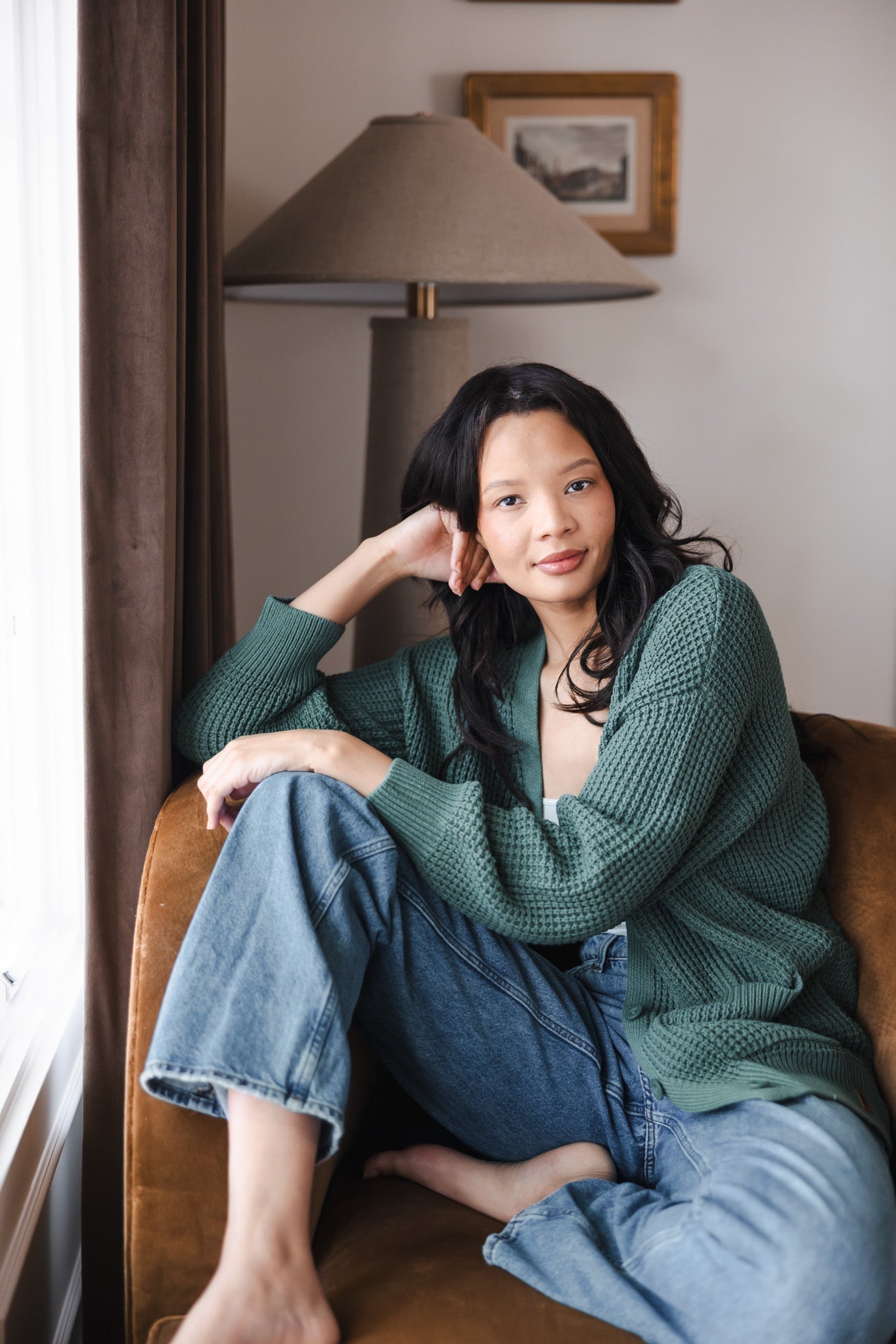 Modal sitting on brown sofa wearing Chunky Knit Women's Waffle Cardigan in Pine over white camisole and blue jeans