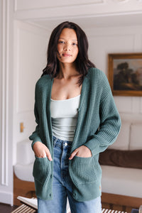 Modal standing in living room wearing Chunky Knit Women's Waffle Cardigan in Pine over white camisole and blue jeans
