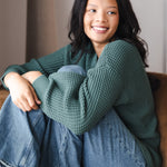 Modal smiling on brown sofa wearing Chunky Knit Women's Waffle Cardigan in Pine over blue jeans