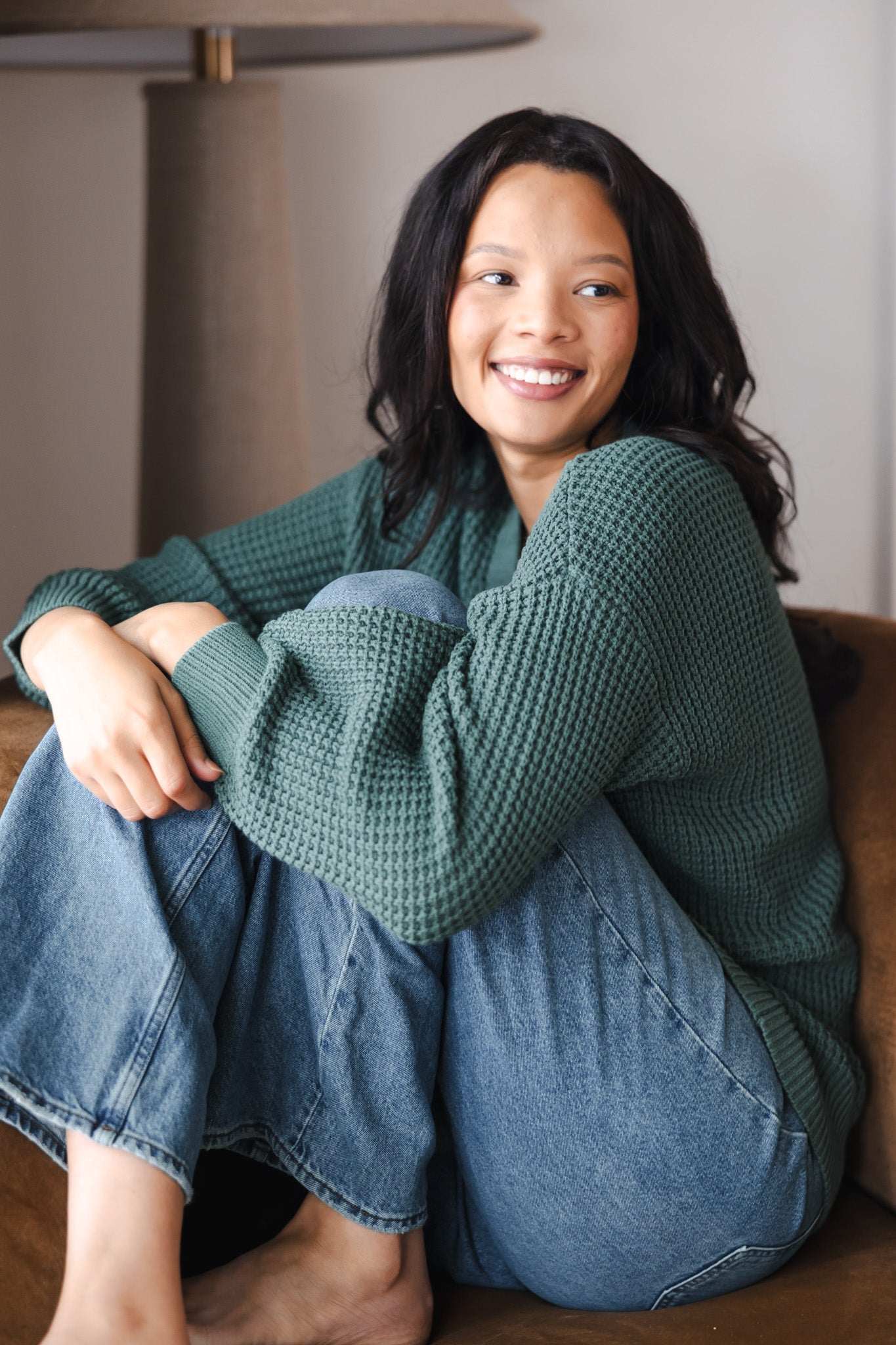 Modal smiling on brown sofa wearing Chunky Knit Women's Waffle Cardigan in Pine over blue jeans