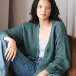 Modal sitting on brown sofa wearing Chunky Knit Women's Waffle Cardigan in Pine over white camisole and blue jeans