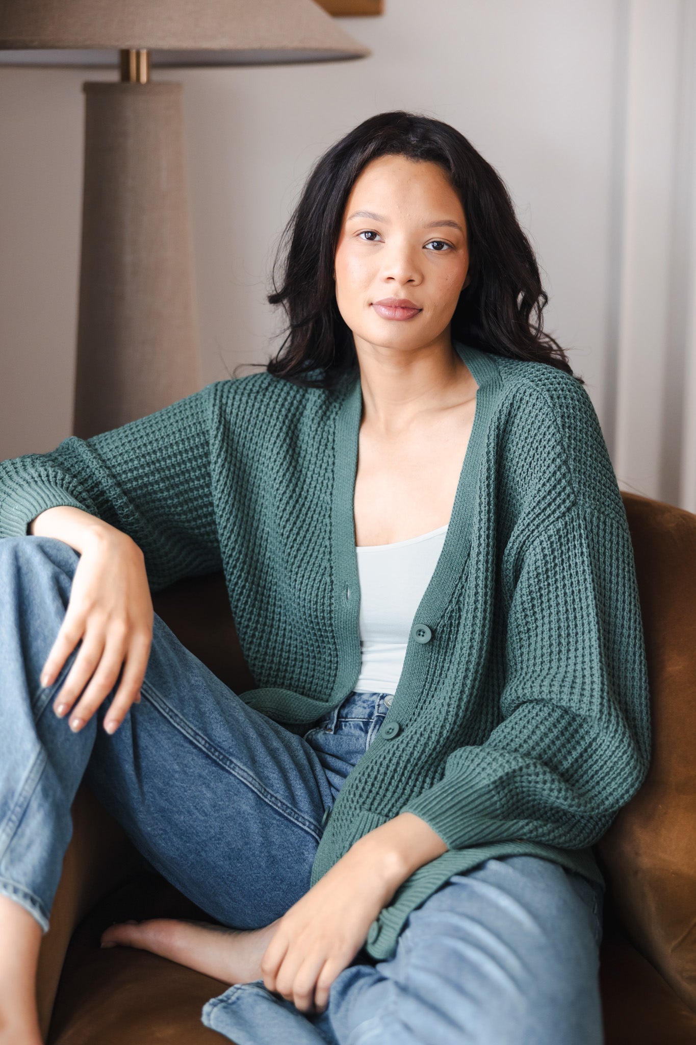 Modal sitting on brown sofa wearing Chunky Knit Women's Waffle Cardigan in Pine over white camisole and blue jeans