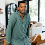 Male model wearing muted sage green button up long sleeve pajamas in a living room