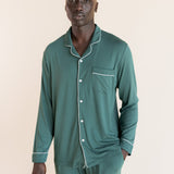 Male model wearing muted sage green button up long sleeve pajamas in a studio setting, hand in pocket