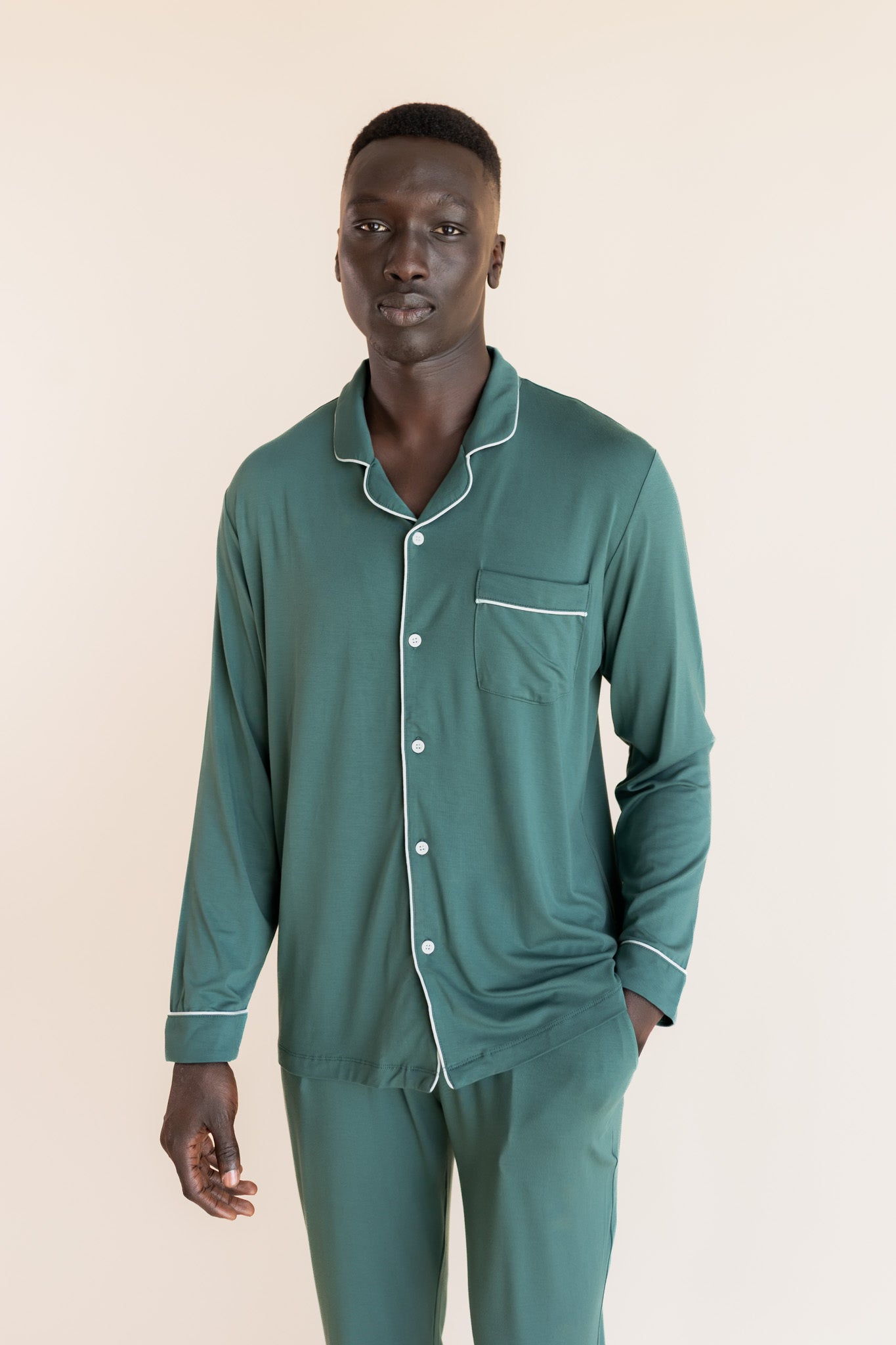 Male model wearing muted sage green button up long sleeve pajamas in a studio setting, hand in pocket