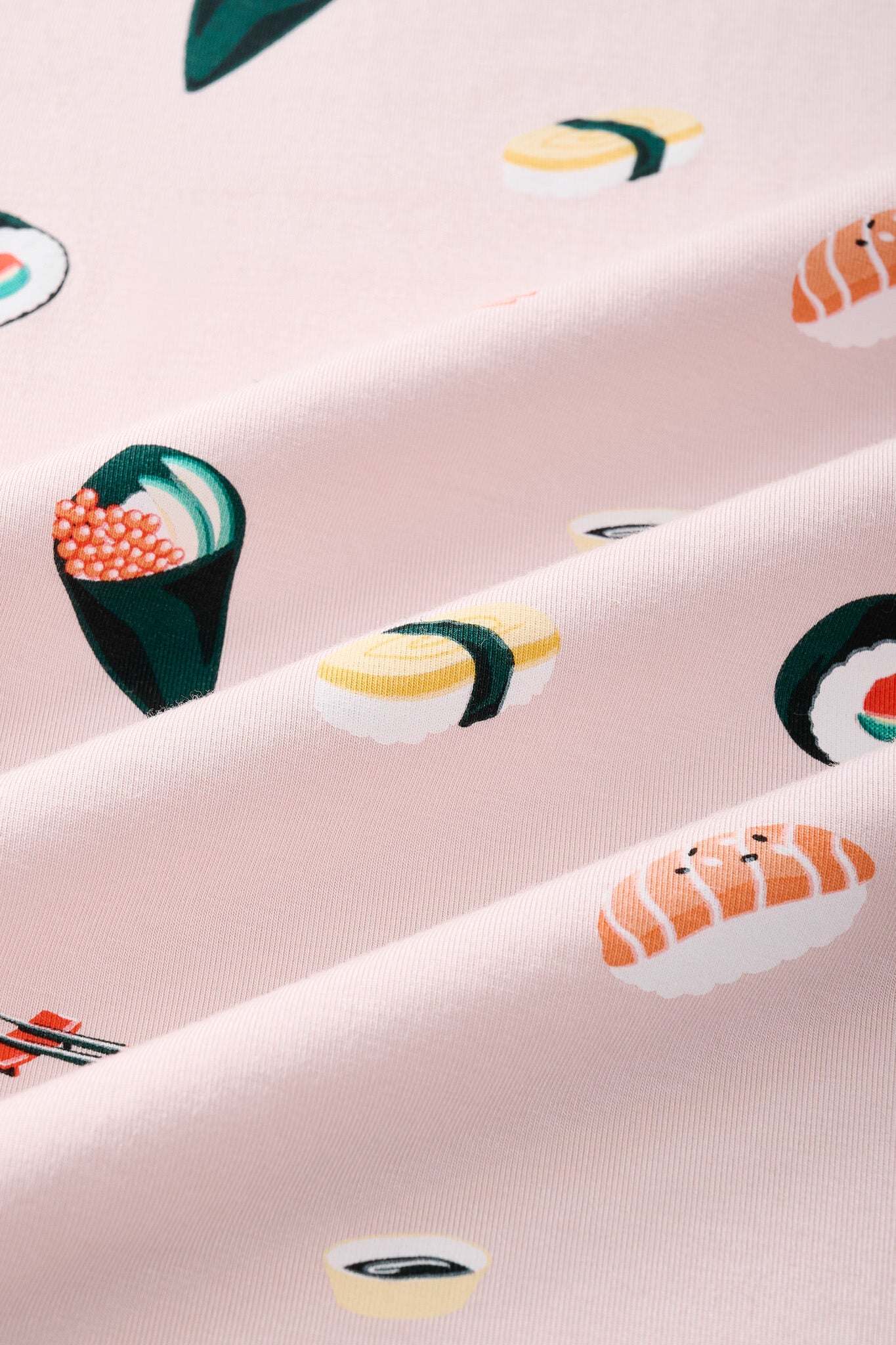 A close-up of a pink fabric with a repeating sushi print, including nigiri, maki, and sashimi motifs in yellow, green, orange, and white.---1615 1