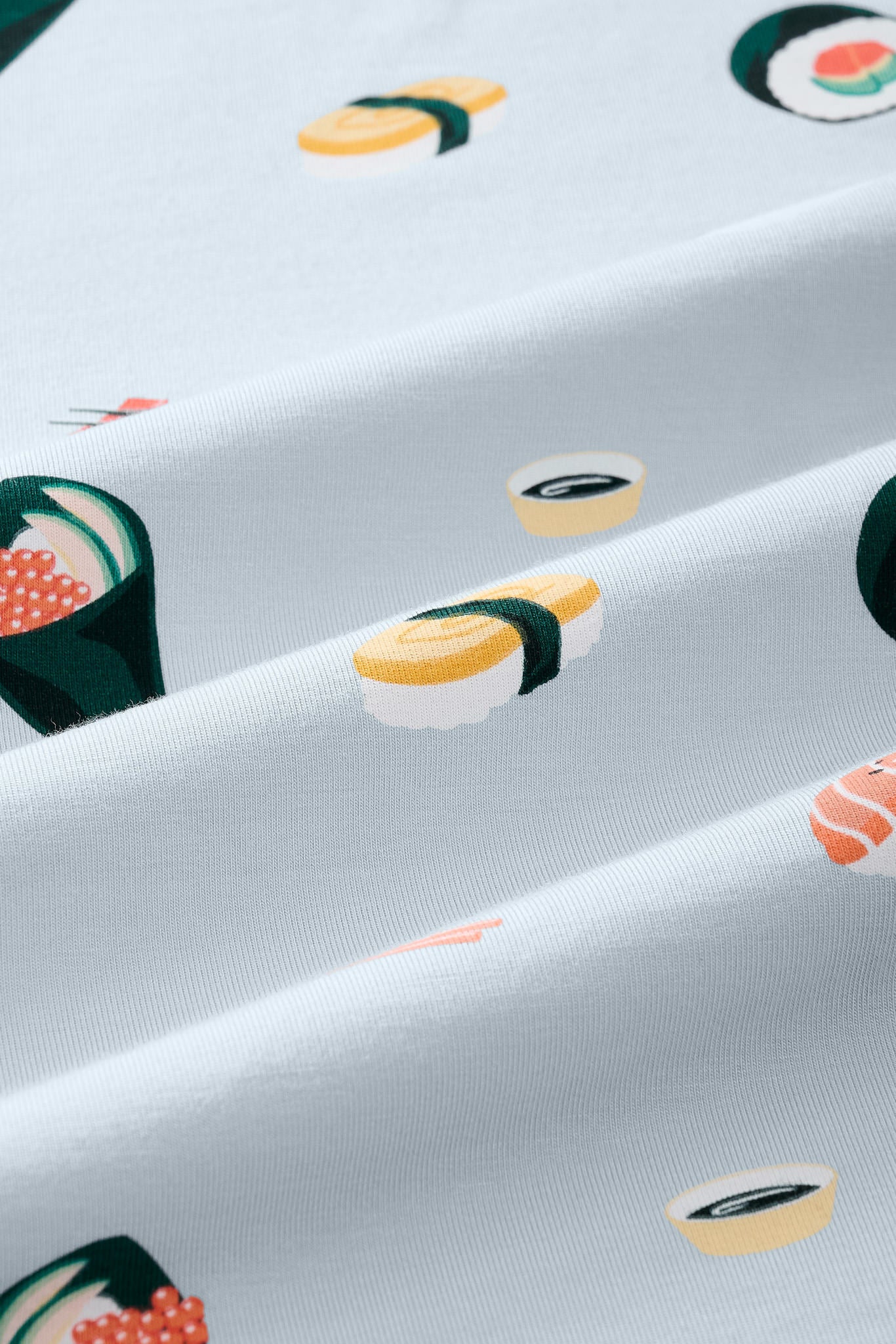 A close-up of a light blue-gray fabric with a repeating sushi print, including nigiri, maki, and sashimi motifs in yellow, green, orange, and white.---1615 1