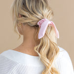Woman with blonde hair styled in a ponytail with a pink scrunchie, wearing a white top against a beige background.