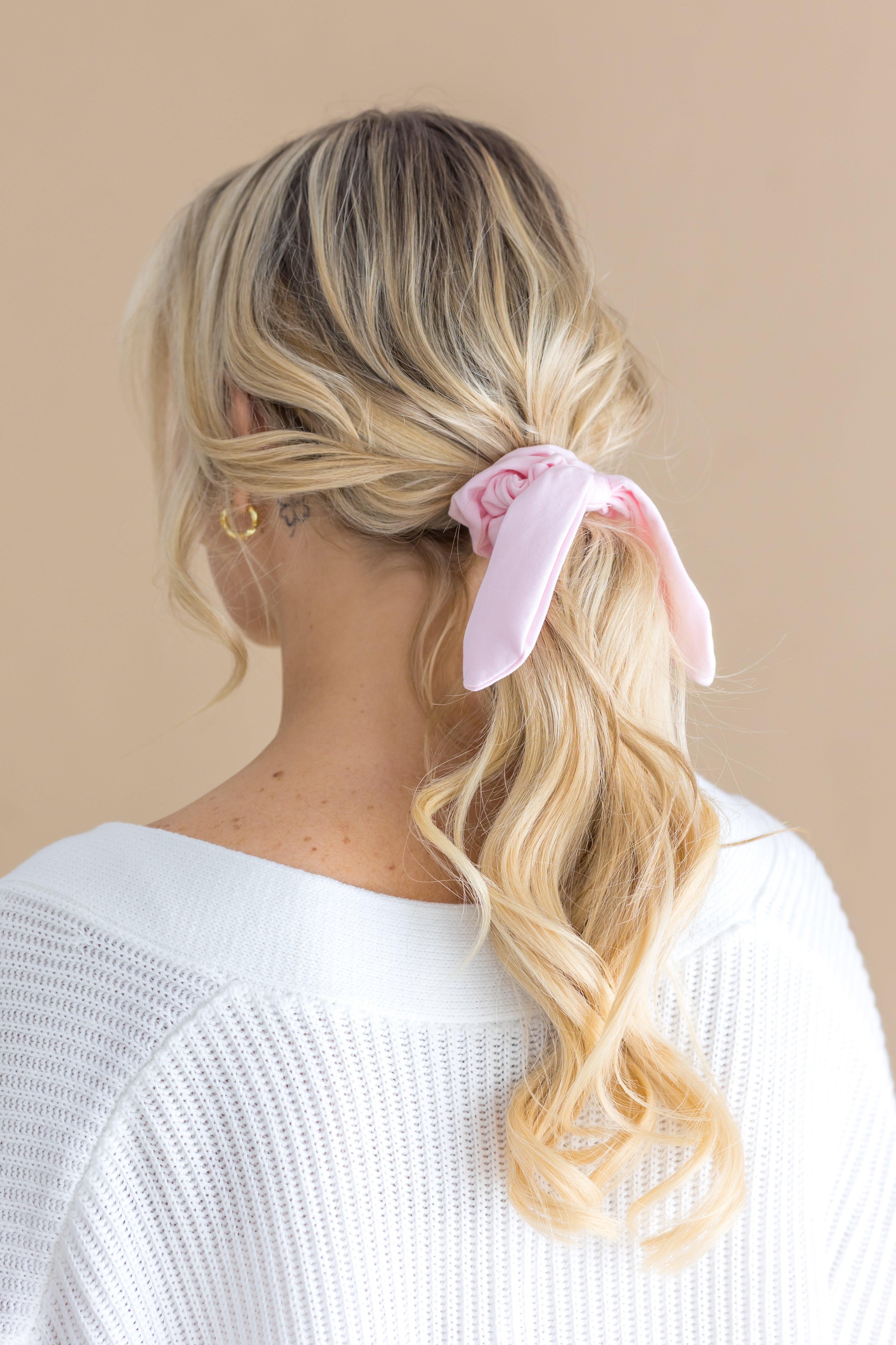 Woman with blonde hair styled in a ponytail with a pink scrunchie, wearing a white top against a beige background.