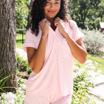 A woman wearing a light pink short-sleeve button-front top and matching shorts stands outdoors in a garden setting with trees and flowers in the background.
