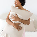 Female kneeling on a bed holding a  pillow with the Bamboo Sateen Pillowcase in Sand Stone on it