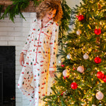 Smiling model looking down next to a Christmas tree while wearing the Kyte Long Sleeve Women's Pajama Set in Santa Sleigh