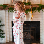 Smiling model standing infront a white brick fireplace while wearing the Kyte Long Sleeve Women's Pajama Set in Santa Sleigh