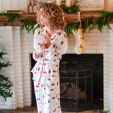Smiling model standing infront a white brick fireplace while wearing the Kyte Long Sleeve Women's Pajama Set in Santa Sleigh