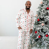 Male model next to a Christmas tree wearing the Kyte Men's Long Sleeve Pajama Set in Santa Sleigh