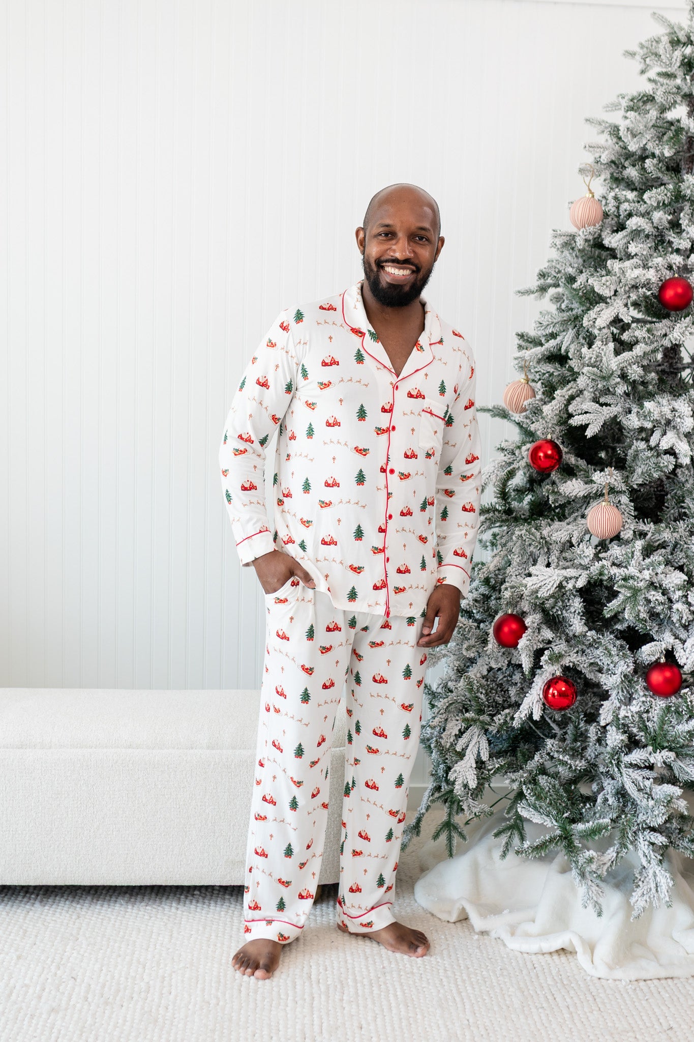 Male model next to a Christmas tree wearing the Kyte Men's Long Sleeve Pajama Set in Santa Sleigh