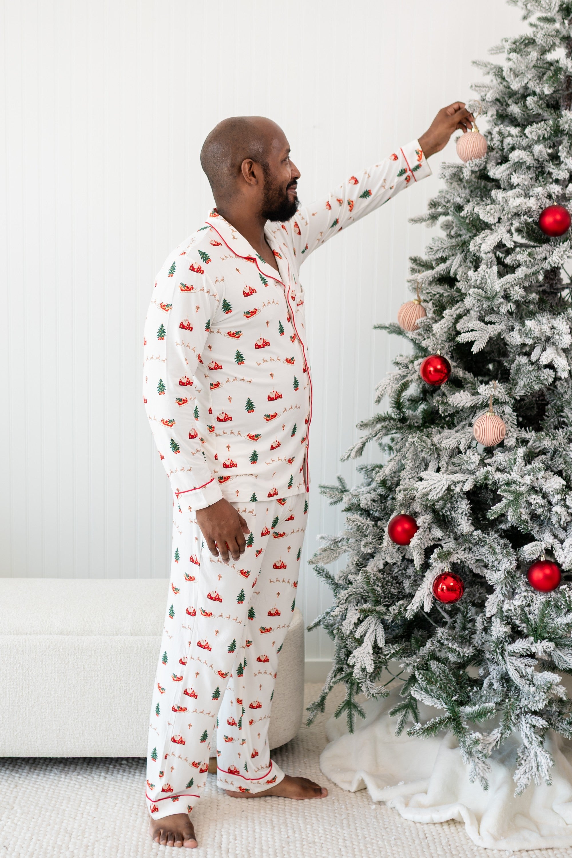 Male model putting an ornament on a Christmas tree while wearing the Men's Long Sleeve Pajama Set in Santa Sleigh