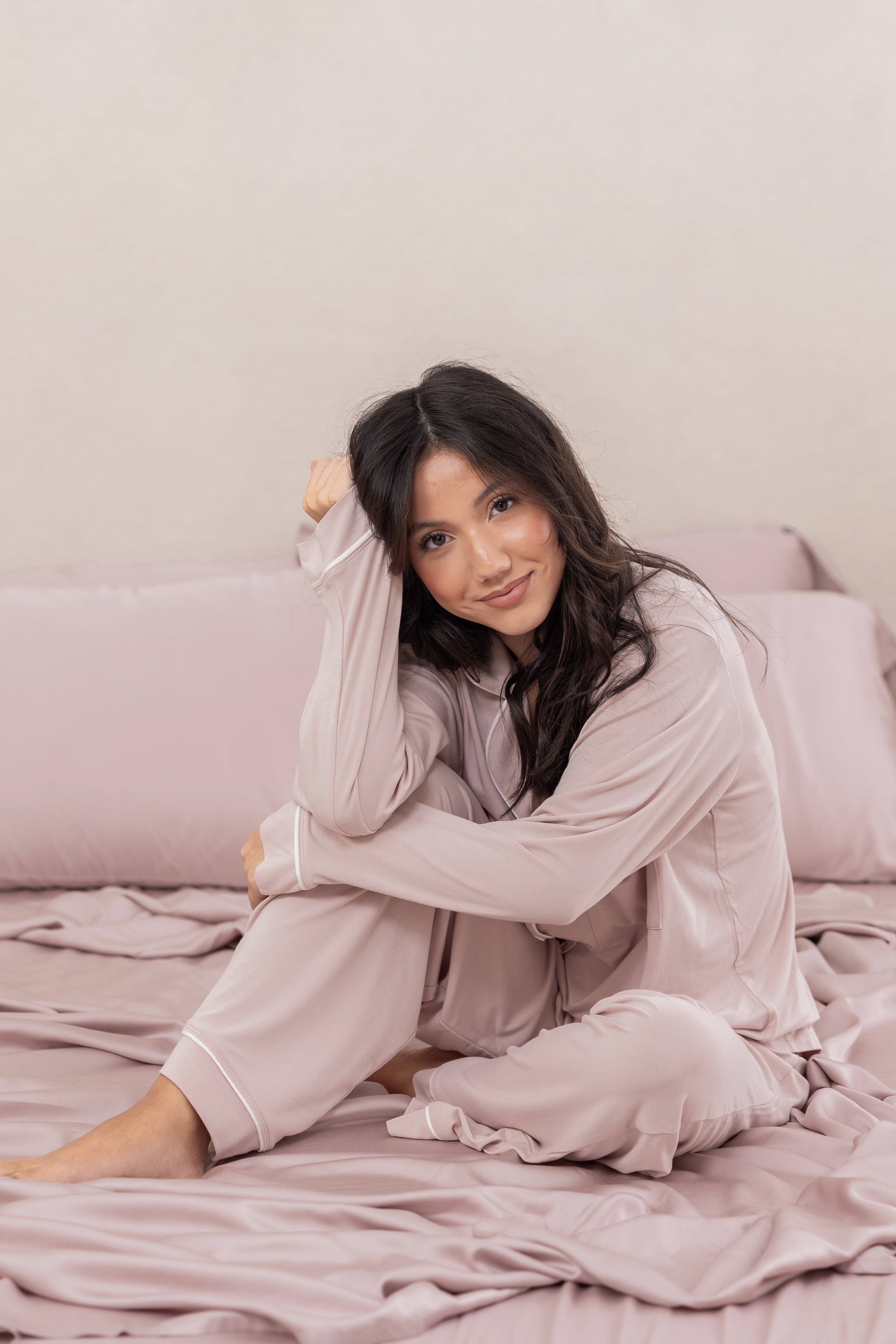 Woman in light pink pajamas sitting on a bed with a neutral background
