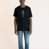 Modal posing in Black Men's Crew Neck Tee with Skeleton print in center over blue jeans on a beige background