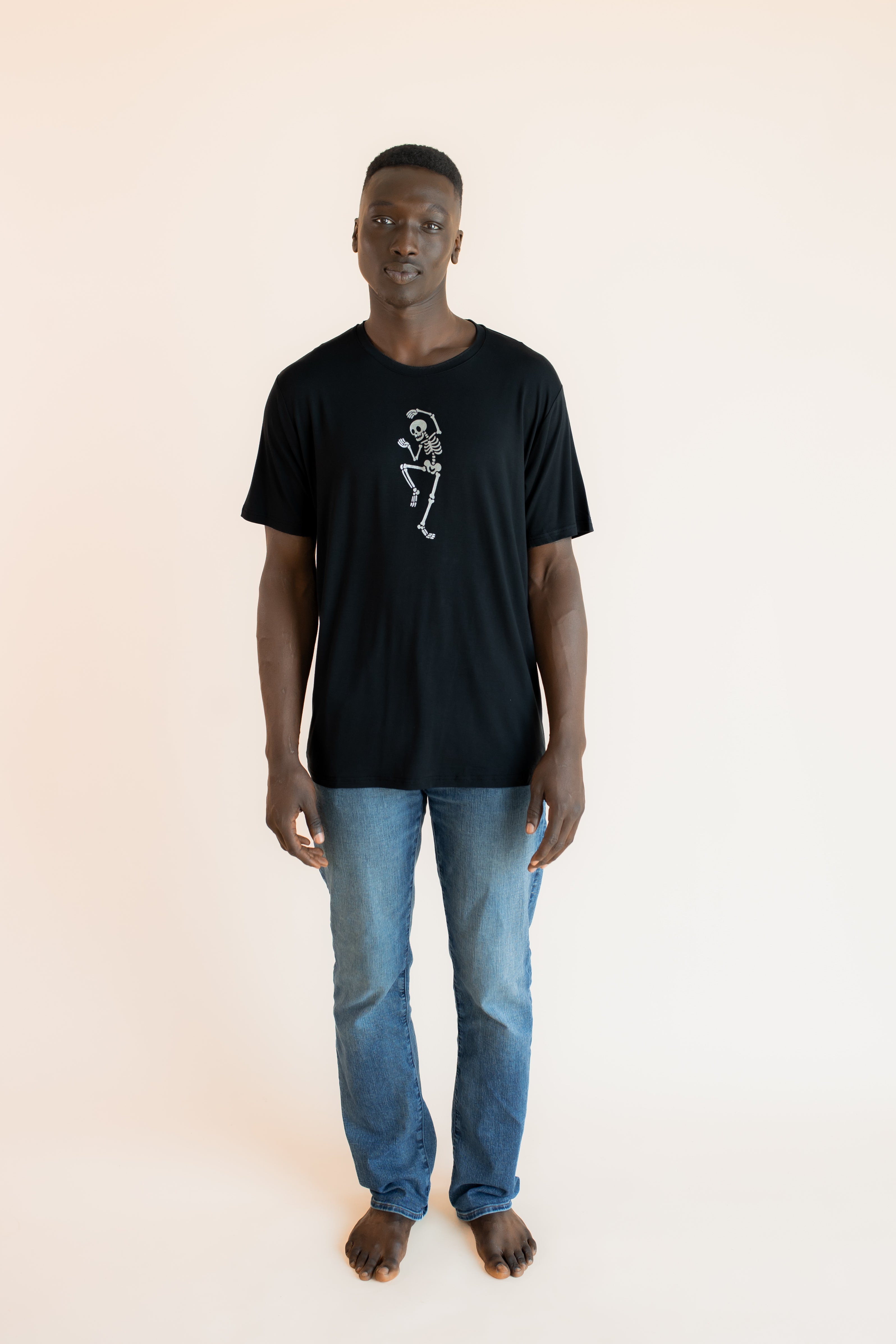 Modal posing in Black Men's Crew Neck Tee with Skeleton print in center over blue jeans on a beige background