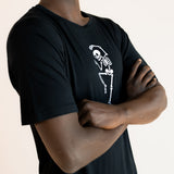 Side pose of Modal wearing Black Men's Crew Neck Tee with Skeleton print in center on a beige background