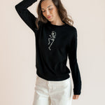 Modal posing in Black Women's Sweatshirt with Skeleton print in the center over white pants on a beige background