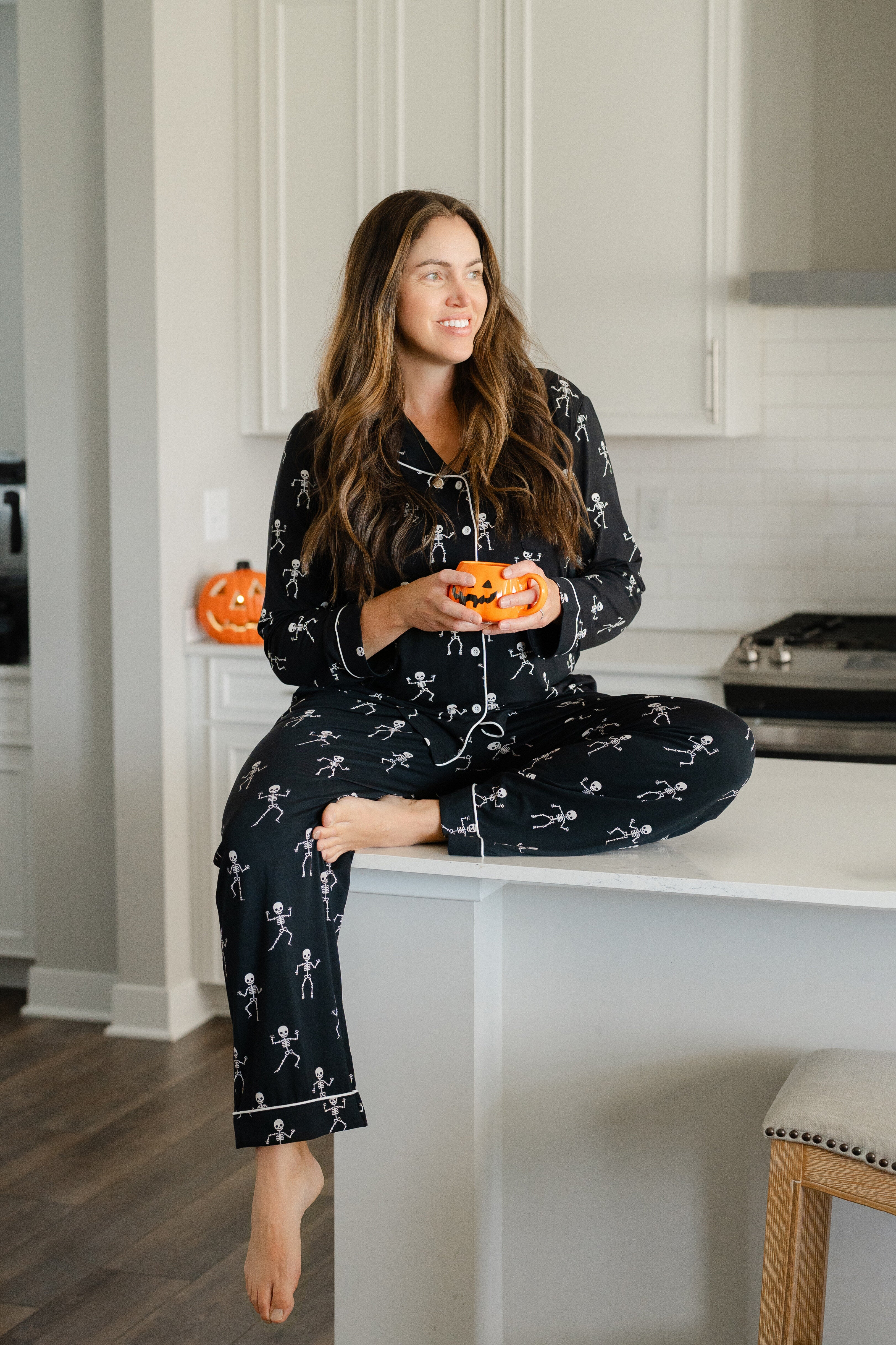 Woman in black skeleton pajamas sitting on a kitchen counter holding small pumpkins.