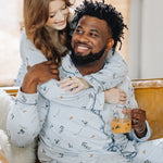 Male sitting on the couch wearing the Men's Long Sleeve Pajama Set in Ski with his wife standing behind him with her arms around her shoulders matching in women's ski pajama set