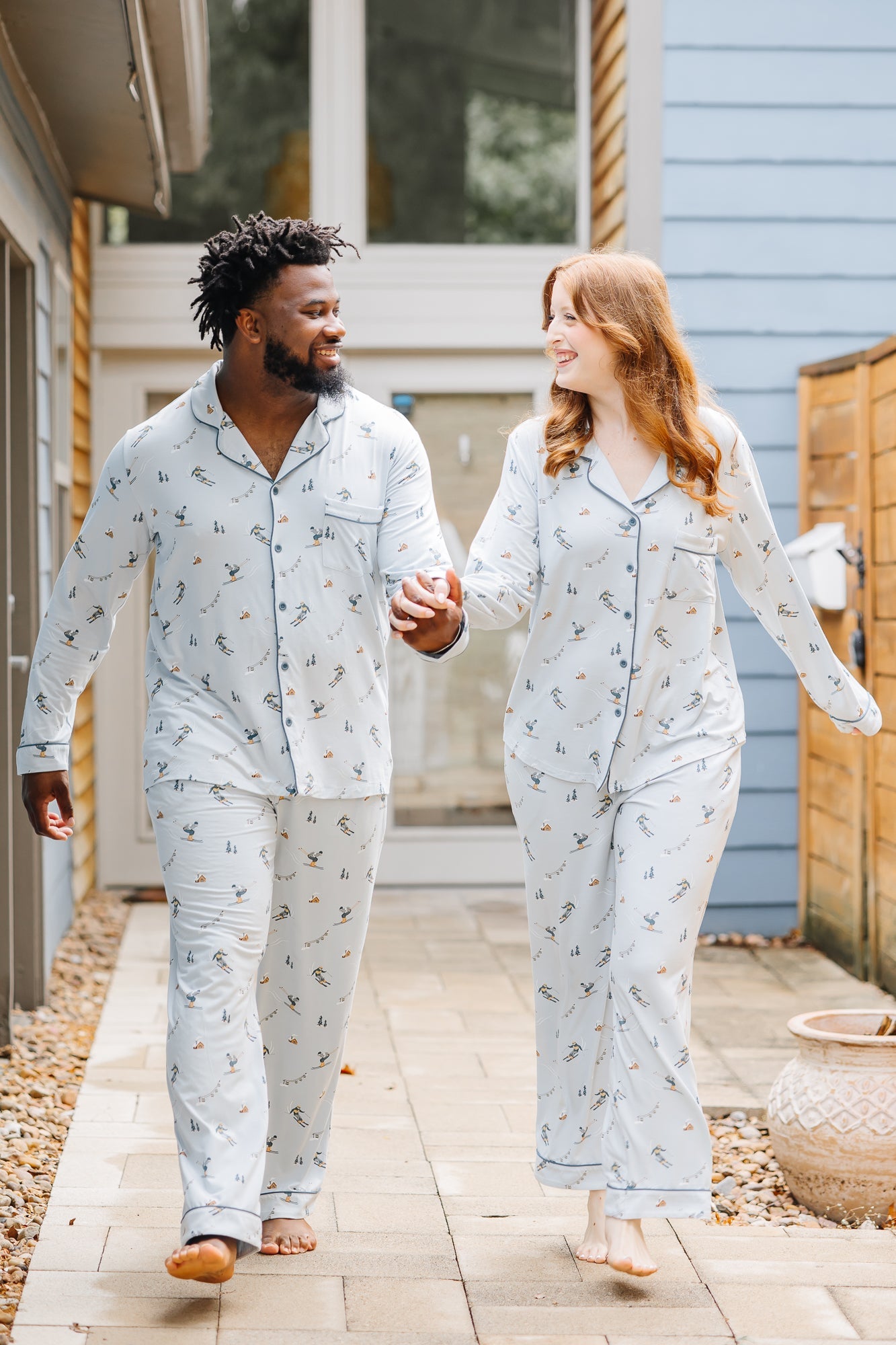 Couple holding hands walking on a tiled sidewalk in front of a house matching in Women's and Men's Long Sleeve Pajama Set in Ski