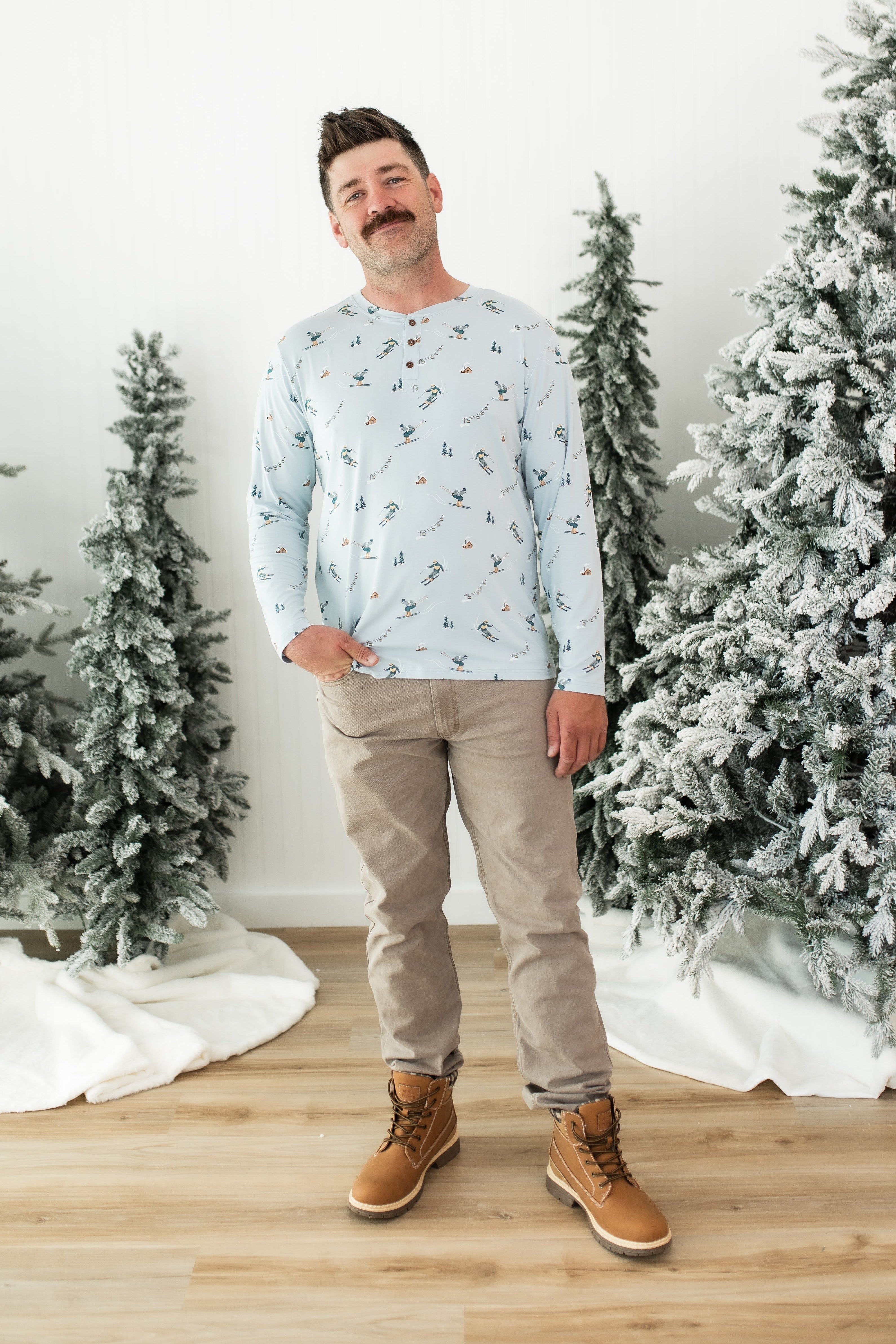 Smiling male modeling the Men's Long Sleeve Henley Top in Ski with khaki colored pants and light brown boots standing between frosted trees