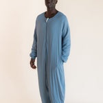 A man wearing a dusty blue long-sleeve zip-up onesie, standing against a plain beige studio background.