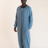 A man wearing a dusty blue long-sleeve zip-up onesie, standing against a plain beige studio background.