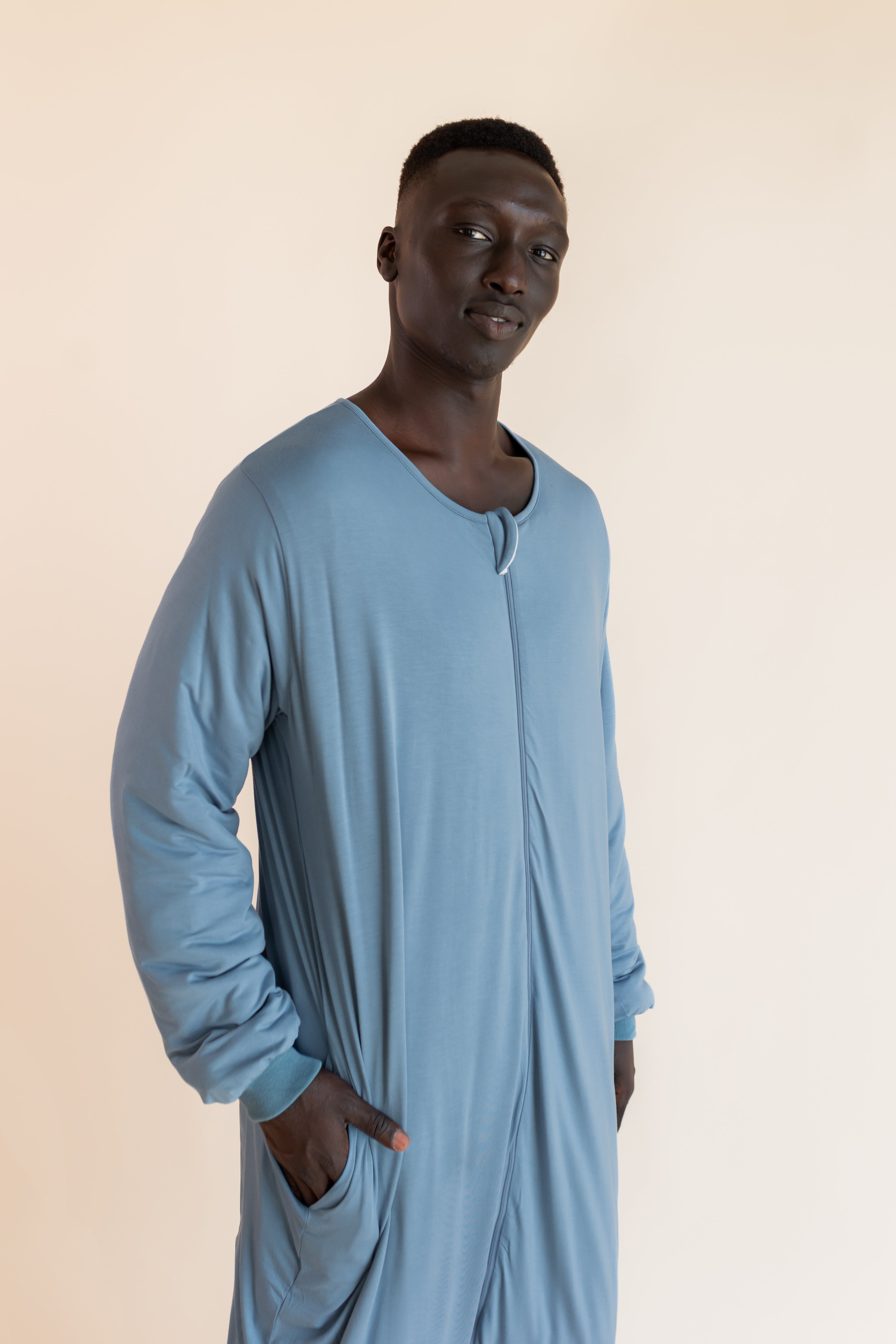 A man wearing a light blue long-sleeve sleep sack, standing against a solid beige background.
