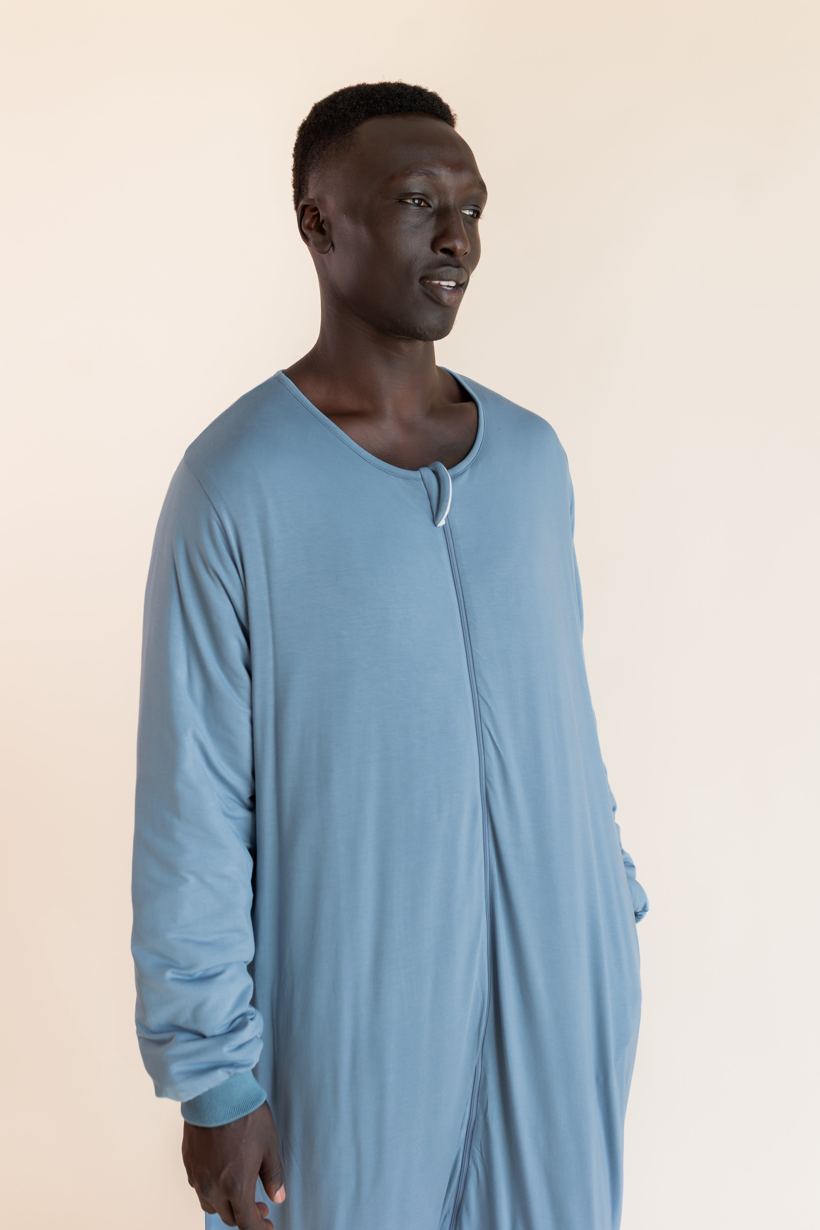 A man wearing a light blue long-sleeve sleep sack, standing against a solid beige background.