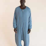 A man wearing a dusty blue, long-sleeve, full-body zip-up harem-style sleep sack against a plain, warm off-white studio background.