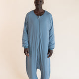 A man wearing a dusty blue, long-sleeve, full-body zip-up harem-style sleep sack against a plain, warm off-white studio background.