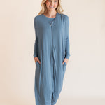 A woman modeling a dusty blue long-sleeve footless sleep sack pajama jumpsuit with side pockets, standing against a solid light beige background.