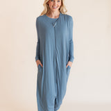 A woman modeling a dusty blue long-sleeve footless sleep sack pajama jumpsuit with side pockets, standing against a solid light beige background.