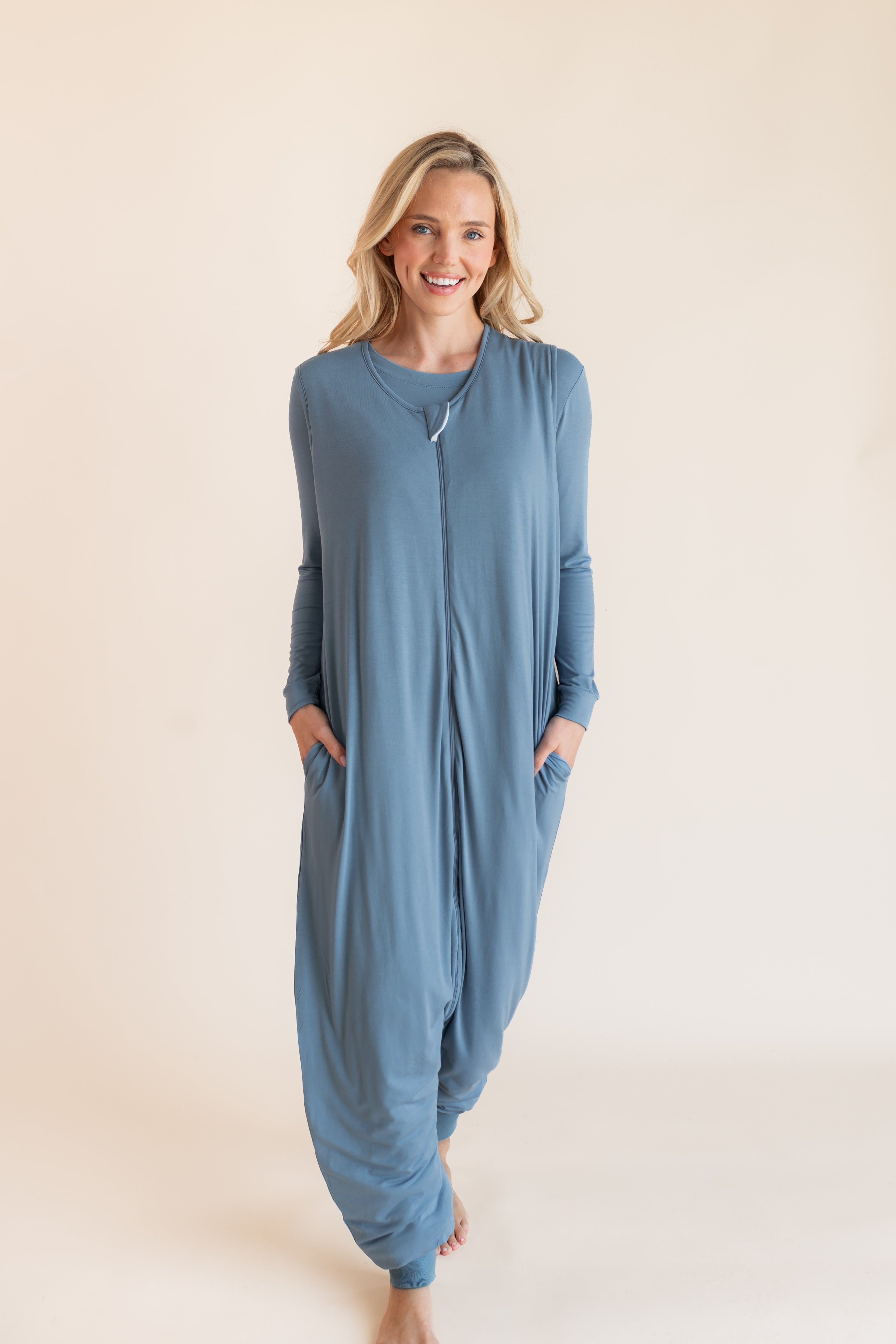 A woman modeling a dusty blue long-sleeve footless sleep sack pajama jumpsuit with side pockets, standing against a solid light beige background.