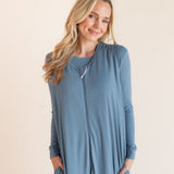 A smiling blonde woman wearing a dusty blue sleeveless-sleeve sleep sack that has pockets with matching shirt underneath