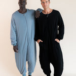 Two men standing barefoot in full-body zip-up onesies against a plain light beige studio background. The man on the left wears a light dusty blue one-piece, and the man on the right wears a black long-sleeve one-piece.