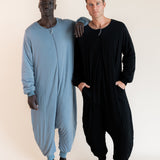 Two men standing barefoot in full-body zip-up onesies against a plain light beige studio background. The man on the left wears a light dusty blue one-piece, and the man on the right wears a black long-sleeve one-piece.