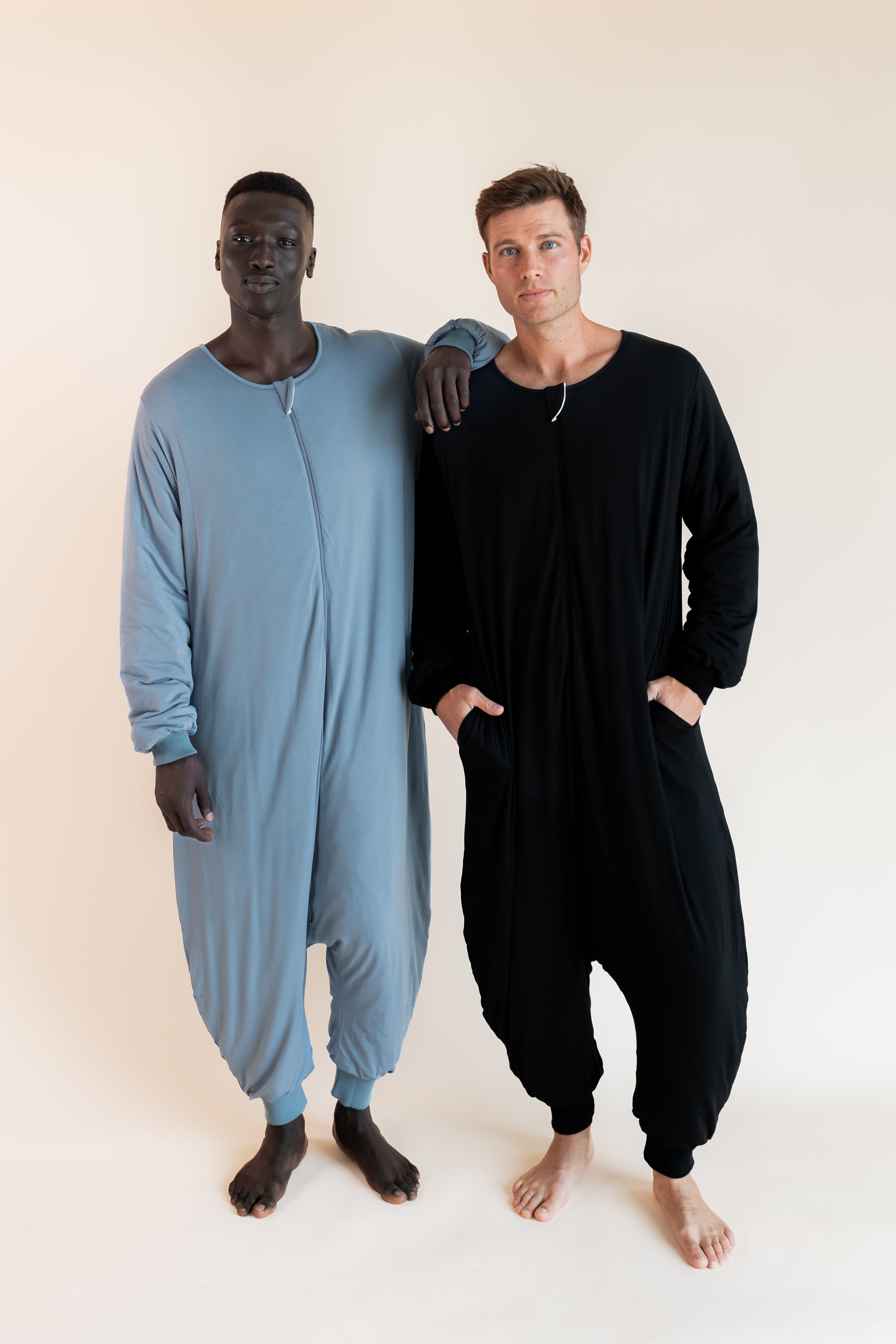 Two men standing barefoot in full-body zip-up onesies against a plain light beige studio background. The man on the left wears a light dusty blue one-piece, and the man on the right wears a black long-sleeve one-piece.